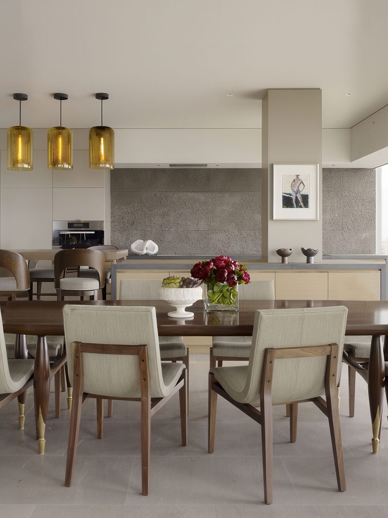 A modern dining room with a wooden table and six beige upholstered chairs. Above the table hang three amber glass pendant lights. The background features a contemporary kitchen with neutral tones, a bouquet of red flowers, and a framed artwork on the wall.