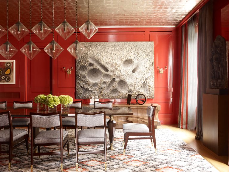 A formal dining room with glossy red walls, a textured abstract art piece, and a rectangular table surrounded by gray upholstered chairs. Glass pendant lights hang above, green flower arrangements sit on the table, and sunlight streams in from tall windows with sheer curtains.