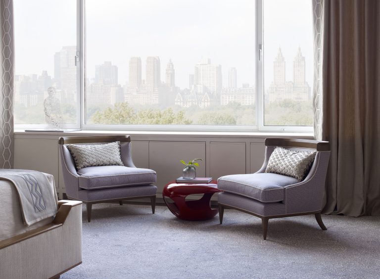 Two armchairs with patterned cushions face each other beside a red side table with a small potted plant. Large window behind them shows a city skyline and trees. Neutral tones dominate the elegant, modern room; part of a bed is visible on the left.