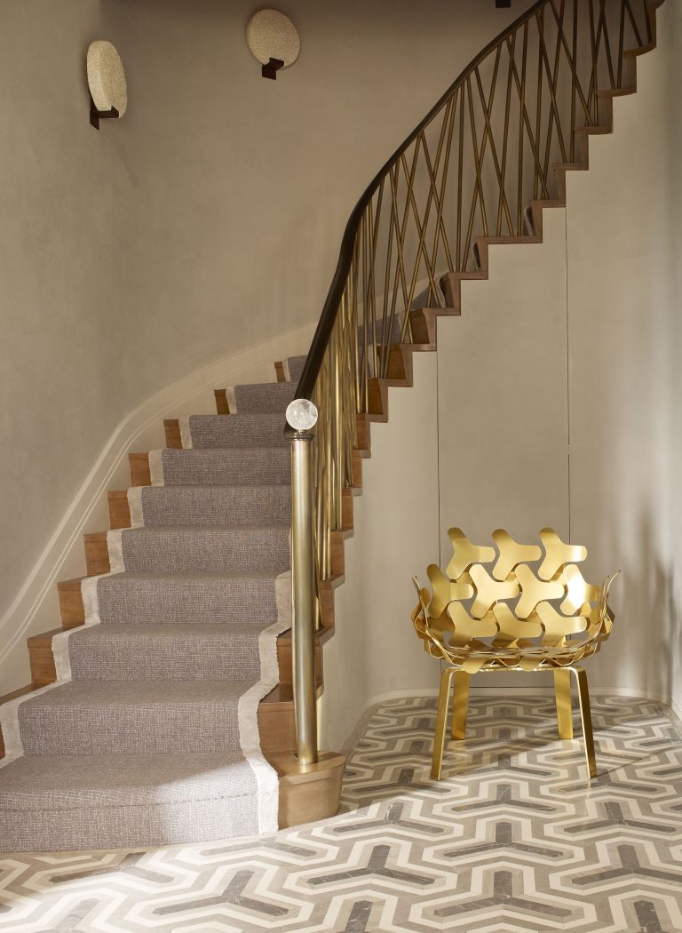 A stylish staircase with a gold handrail and gray carpet runner curves upward. The floor features a geometric patterned tile in beige and taupe. A sculptural gold chair with interlocking shapes sits next to the stairs, and two round wall lights are mounted above.