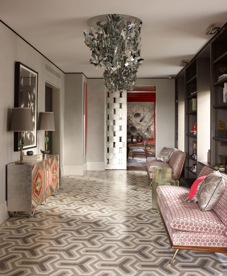 A stylish living room with patterned geometric floor tiles, two patterned sofas, a modern chandelier with metallic leaves, a console table with lamps and geometric doors, bookshelves, and a view into a red-painted adjoining room. Natural light fills the space.