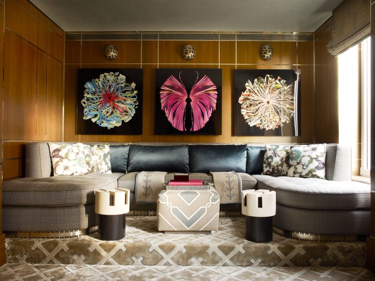 A modern living room with a gray curved sectional sofa, patterned pillows, two small round white tables, a geometric ottoman, and a beige rug. Three large, colorful abstract floral artworks hang on wood-paneled walls above the sofa.