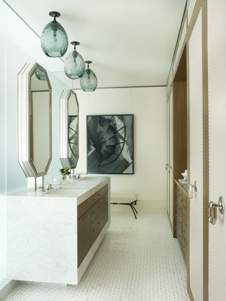 A modern bathroom with two octagonal mirrors above a marble double sink, dark wood cabinets, and three green glass pendant lights. Geometric art hangs on a white wall near a bench; the floor has a white hexagonal tile pattern.
