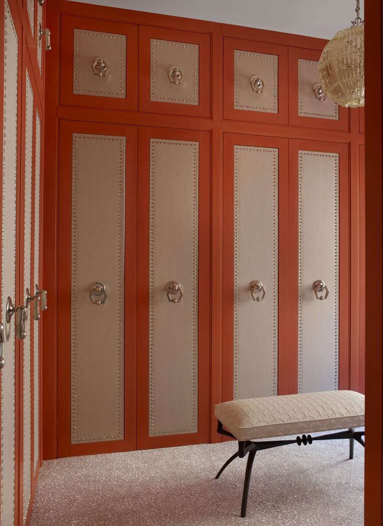 A room with tall, paneled wardrobes in burnt orange and beige, decorated with ornate circular handles. The floor is covered in a light, speckled pattern. A quilted cream bench with a dark, wrought iron base sits in front. A glass pendant light hangs above.