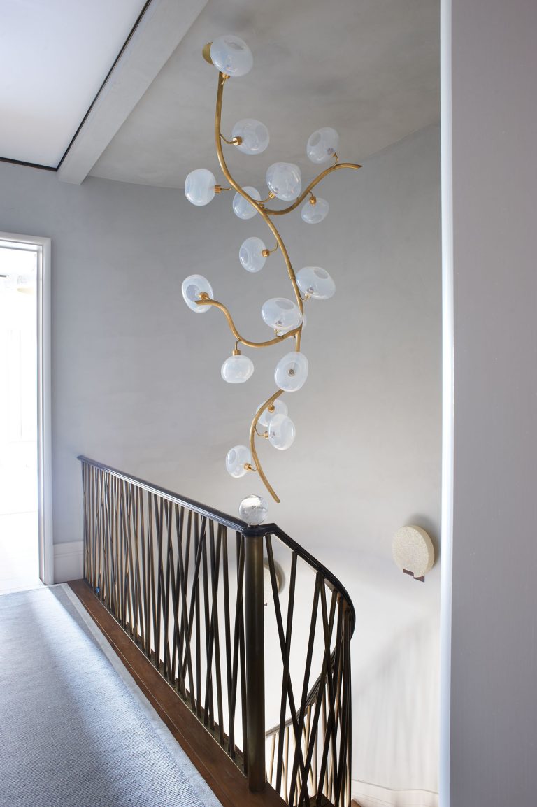 A modern staircase features a sculptural gold chandelier with delicate, branching arms and translucent glass orbs. The metal stair railing has an abstract, angular pattern. Soft natural light enters from an open doorway, highlighting the clean, minimalist decor.