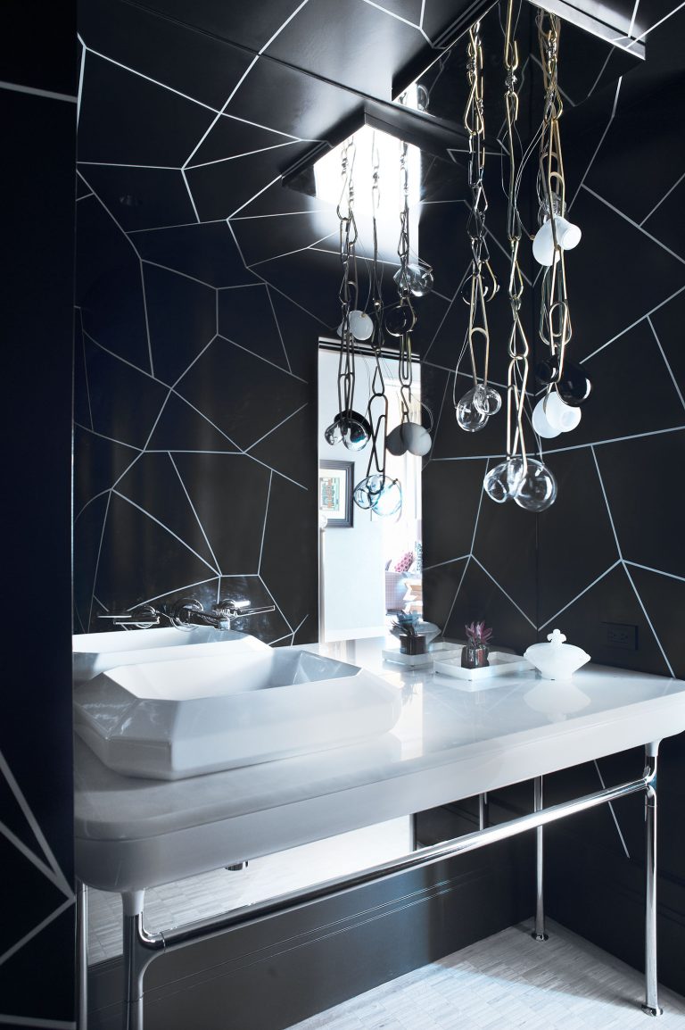 A modern bathroom with black geometric wall tiles, a white rectangular sink on a glossy white countertop, and multiple hanging light bulbs with exposed cords. A small potted plant and minimalist decor sit on the counter under a skylight.
