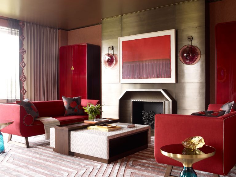 A modern living room with two red sofas, red cabinets, a geometric fireplace, and a red abstract painting above it. The decor includes a gold side table, a patterned rug, and neutral curtains with red accents. The room has a warm, sophisticated atmosphere.