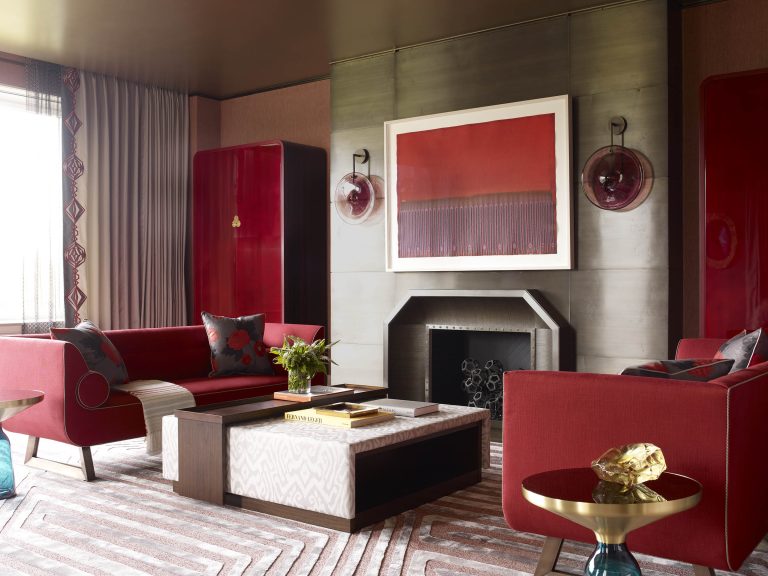 A modern living room with bold red sofas, a central coffee table, gold and wood side tables, and geometric-patterned rug. The fireplace is framed by brass panels, with a large red abstract painting and two round wall sconces above it. Decor is elegant and contemporary.