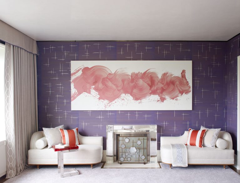 A modern living room with lavender walls patterned with silver crosses, two white sofas with orange and gray pillows, a glass coffee table, a mirrored fireplace, and a large abstract pink artwork above the mantel. Light enters through tall draped windows.