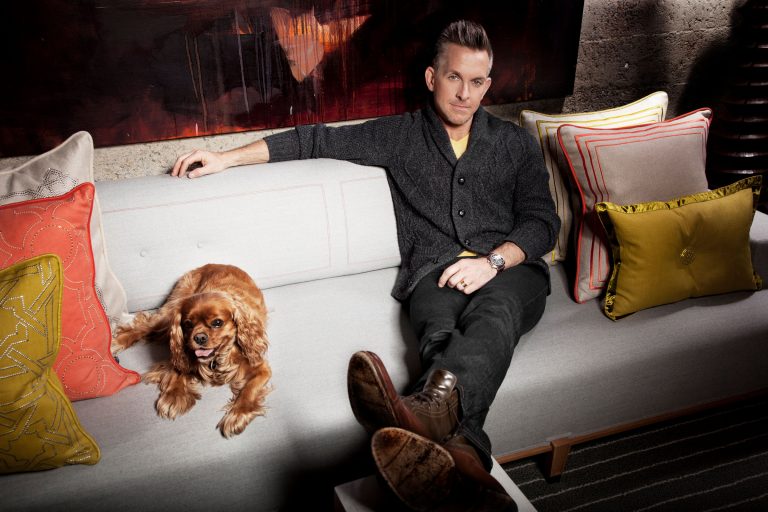 A man in dark clothes sits relaxed on a modern light-gray sofa beside a brown, long-haired dog. Decorative pillows with patterns and textures surround them. Behind is an abstract painting and a textured wall. The mood is cozy and stylish.