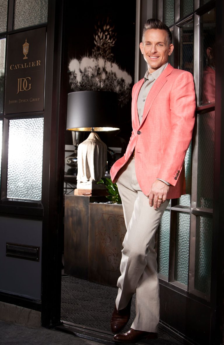 A smiling man in a coral blazer, light shirt, and beige trousers leans casually against a doorway. Behind him is a stylish interior with a dark lamp, decorative plants, and a sign reading Cavalier JDG Jeffrey Design Group.