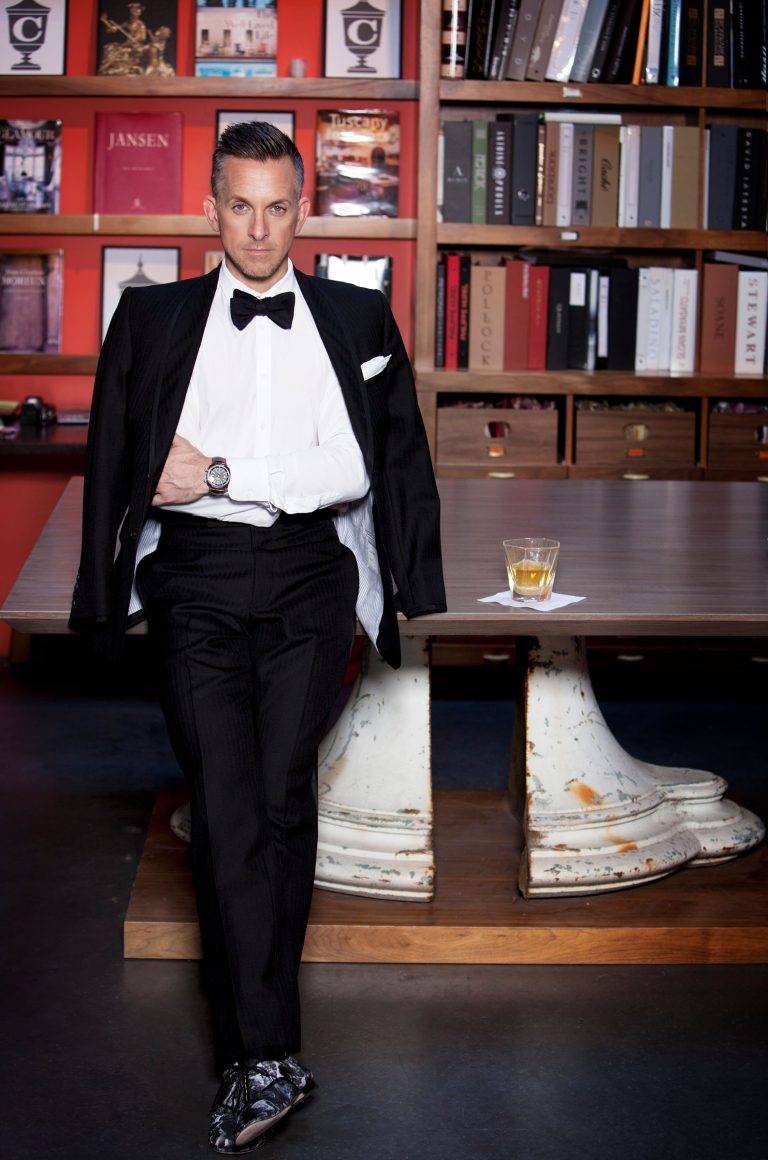 A man in a tuxedo stands confidently, one hand in his pocket, jacket draped over his shoulders. He is in front of a wooden table with a drink on it and bookshelves filled with books and decorative items in the background. The setting appears stylish and sophisticated.