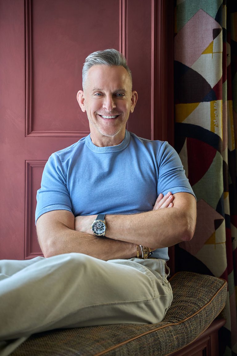 A smiling middle-aged man with short gray hair sits with arms crossed, wearing a light blue T-shirt and light-colored pants. He has a watch and bracelets on his left wrist. He sits on a cushioned bench against a maroon wall and abstract-patterned curtains.