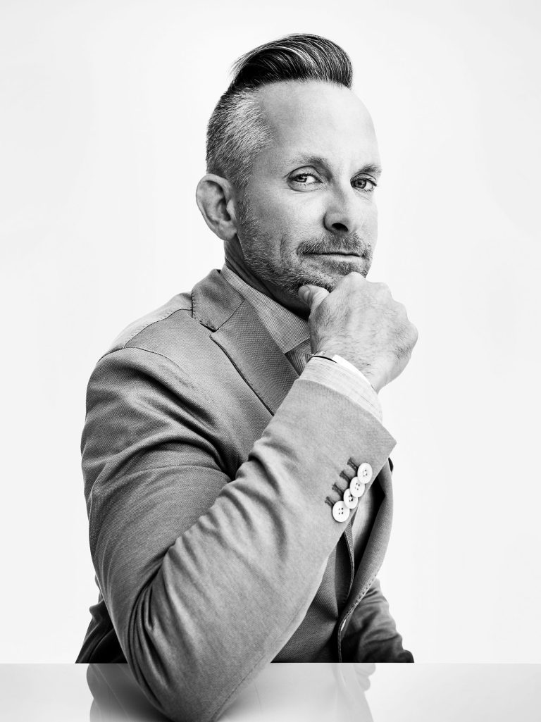 A man in a light-colored suit sits in profile against a white background, looking confidently at the camera. He rests his chin on his hand, has short, styled hair, a trimmed beard, and wears a slight, knowing smile.