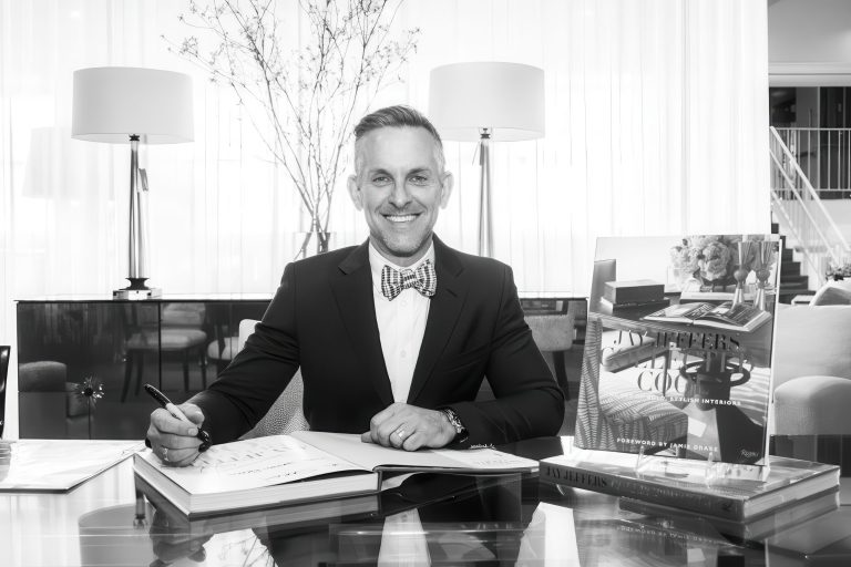 A man in a suit and bow tie smiles while sitting at a desk with open books, holding a pen. He is in a bright, modern office with large windows, two lamps, and decorative branches in the background. A stack of books is displayed next to him.