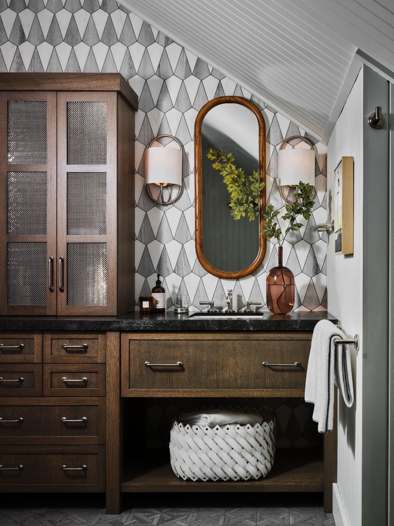 A modern bathroom features dark wood cabinetry, a black countertop, and an oval mirror with a wooden frame. Two sconces flank the mirror. A geometric black-and-white tile wall and a decorative vase with greenery complete the stylish space.