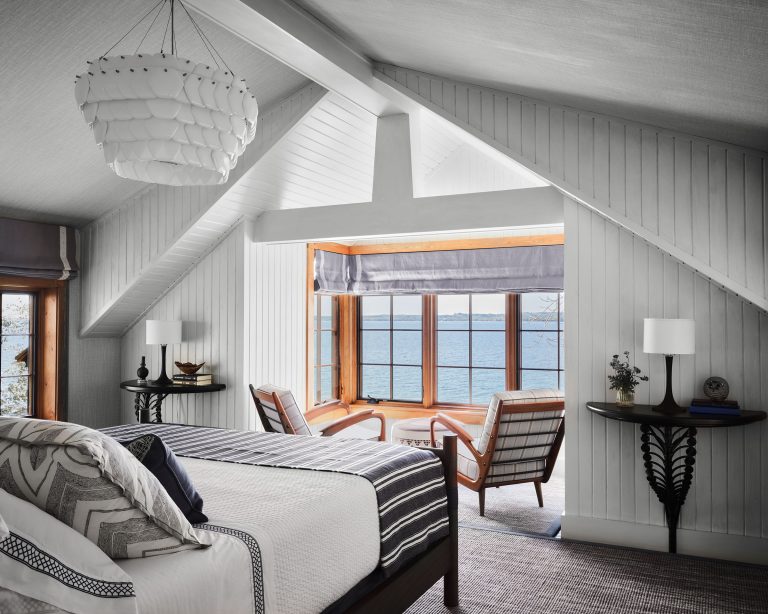 Bright bedroom with white paneled walls and sloped ceiling, large window seat with two armchairs overlooking water, modern black and white decor, bed with patterned pillows and blankets, round tables with lamps, and a sculptural white chandelier.