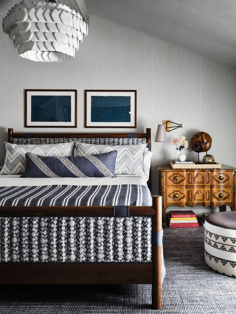 A cozy bedroom features a wood-framed bed with blue and white patterned bedding and pillows. Above the bed are two abstract blue artworks. A vintage wooden dresser with decor sits to the right, under a sloped ceiling and a modern white pendant light.