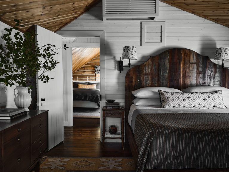 A cozy bedroom with a rustic, dark wood bed and headboard, striped bedding, patterned pillows, white shiplap walls, sloped wood ceiling, nightstand, black dresser, leafy plant in a white vase, and an open door revealing another room with a bed.