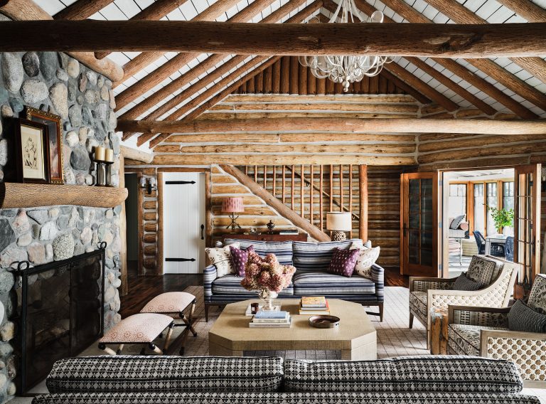 A cozy log cabin living room with exposed wooden beams, a stone fireplace, striped and patterned sofas, a large coffee table, floral arrangement, patterned benches, and open doors leading to a sunroom with wicker seating. Natural light fills the rustic space.