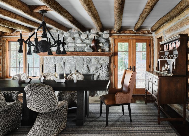 A rustic dining room with a stone fireplace, exposed log beams, a black table with six woven chairs and one brown leather chair, black chandelier, striped rug, wooden hutch, and glass doors letting in natural light.