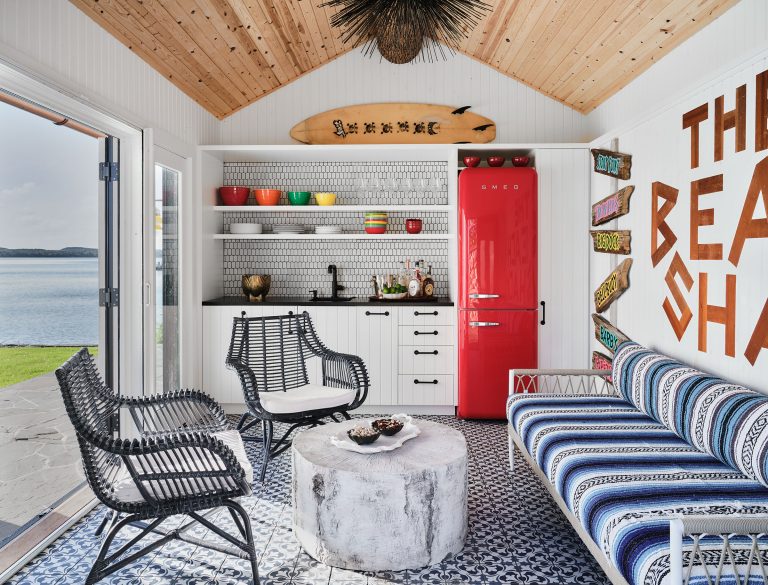 A cozy, beach-themed room with blue-and-white patterned floor tiles, a striped sofa, black wicker chairs, a white round coffee table, open shelves with colorful dishes, a retro red fridge, surfboard decor, “THE BEACH SHACK” wall art, and wide open doors to a water view.