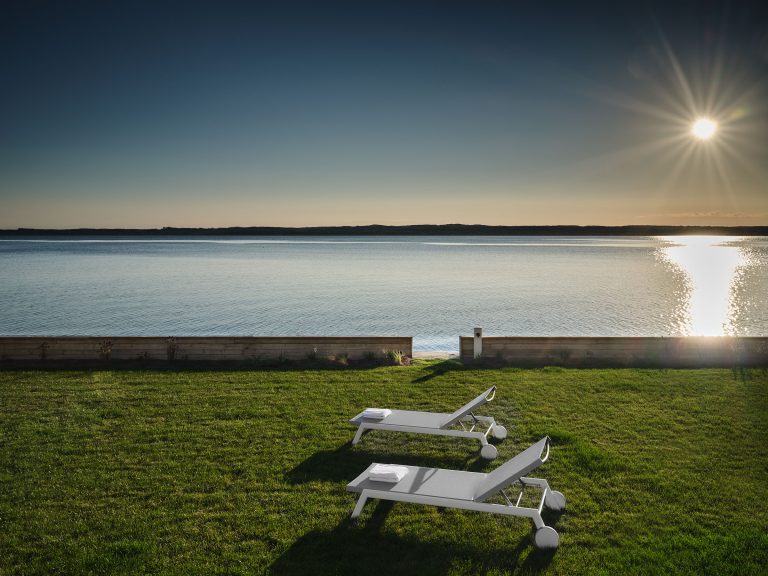 Two white lounge chairs sit on a green lawn overlooking a calm body of water at sunset. The sun is low in the sky, casting a golden reflection on the water. The scene is peaceful, with a clear sky and a wooden barrier separating the lawn from the water.