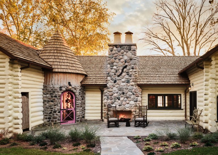A charming log cottage with a turret, stone chimney, and shingle roof sits surrounded by autumn trees. A pink Dutch door opens to the courtyard, where two rocking chairs face an outdoor fireplace on a stone patio, bordered by neat landscaping.
