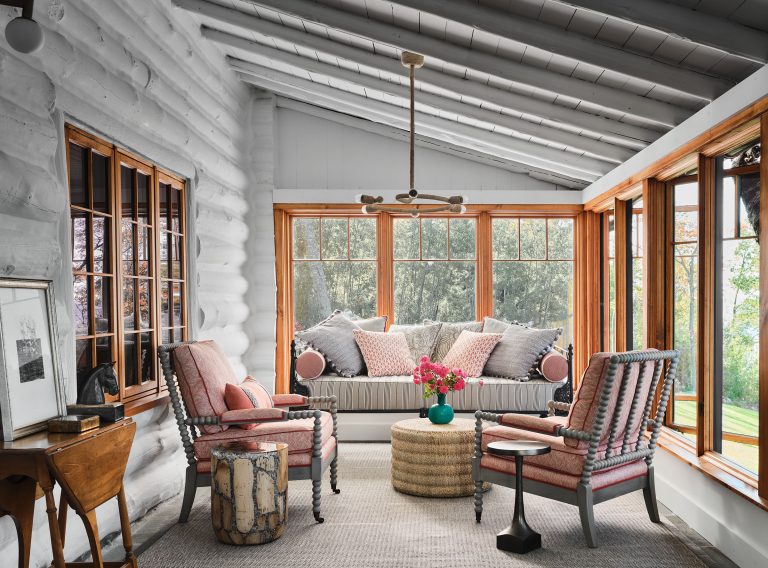A cozy sunroom with whitewashed log walls and large windows on two sides. There’s a built-in daybed with many pillows, two pink upholstered armchairs, a woven round ottoman, a small round table with flowers, and a side table with art and decor pieces.