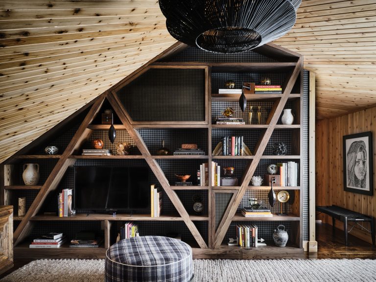 A cozy attic room features slanted wood-paneled ceilings and walls, a large triangular shelving unit filled with books, vases, and decor, a wall-mounted TV, a round plaid ottoman, and a wicker pendant light. A framed portrait hangs on the right wall above a black bench.