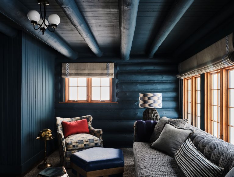 A cozy log cabin room with dark blue log walls and ceiling beams. There’s a gray sofa with patterned pillows, a plaid armchair with a red pillow, a blue ottoman, a brass lamp, and large windows with beige Roman shades letting in natural light.