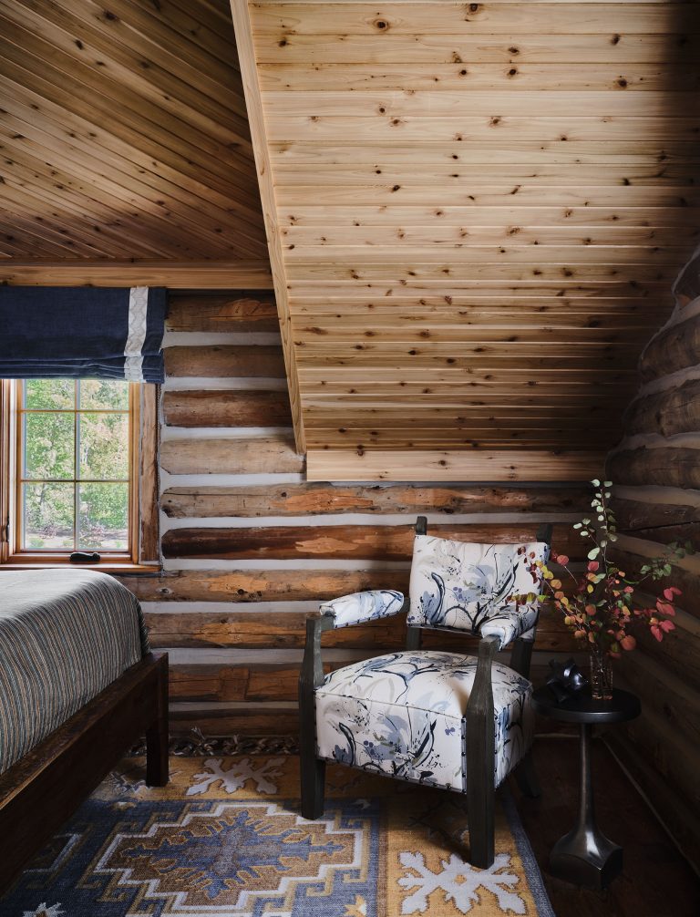 A cozy log cabin bedroom corner with a sloped wood ceiling, log walls, a window with blue curtains, part of a bed, a patterned rug, a floral armchair, and a small black side table holding a vase with branches and a dark cup. Natural light fills the space.