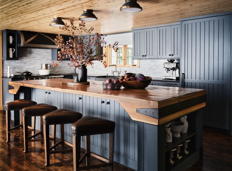 A modern rustic kitchen with blue-gray cabinets, wooden counters, and a large island. Four brown leather stools line the island. A black vase with branches and a wooden bowl of fruit sit on the counter. The room has wood-paneled ceiling and dark wood floors.