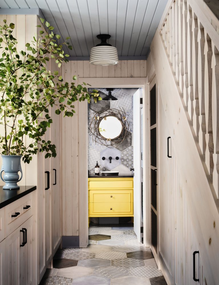 A narrow hallway with light wood walls and cabinetry leads to a bathroom with a yellow vanity. A round mirror with a twig frame hangs above the sink. A leafy branch in a blue vase sits on the left. The patterned floor and ceiling feature geometric tiles and paneling.