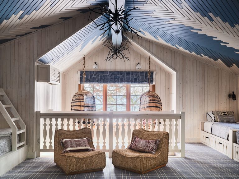 A cozy loft with woven chairs, patterned pillows, and a balustrade overlooks a large window with blue-patterned curtains. The space features light wood paneling, a chevron blue and white ceiling, hanging wicker lights, and beds with plaid bedding.