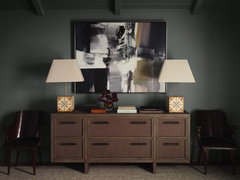 A wooden sideboard with six drawers sits between two dark leather chairs. On the sideboard are two geometric lamps with white shades, a vase with dark flowers, and stacked books. Above hangs an abstract black, white, and gray painting on a dark wall.