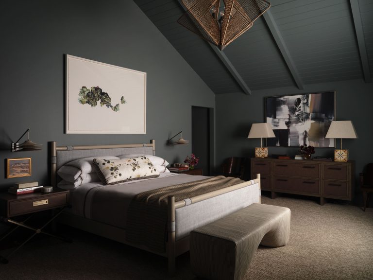 A modern bedroom with dark walls and ceiling, a large bed with gray and white bedding, two nightstands, reading lights, and a bench at the foot. A dresser holds two lamps and art; contemporary art hangs above it. Soft lighting creates a cozy ambiance.