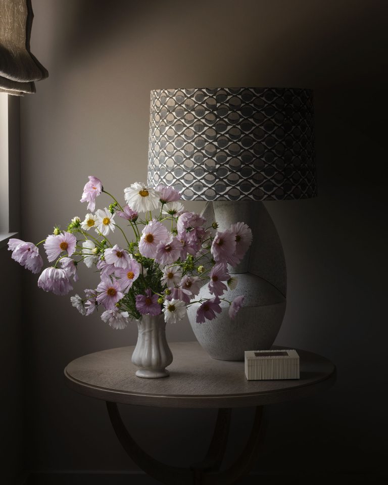 A small round table by a window holds a white vase of pink and white wildflowers, a patterned gray lamp, and a box of matches. Soft natural light highlights the flowers, casting gentle shadows in the dimly lit room.