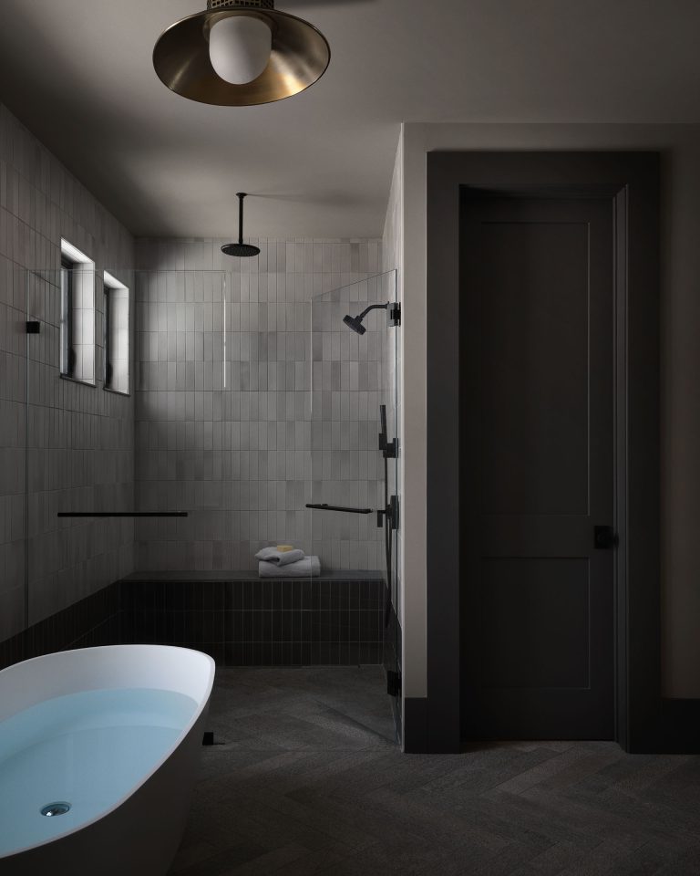 A modern bathroom with dark gray walls and tiles features a freestanding white bathtub filled with water, a walk-in shower with black fixtures, two small windows, folded white towels, and a black door. The lighting is soft and ambient.