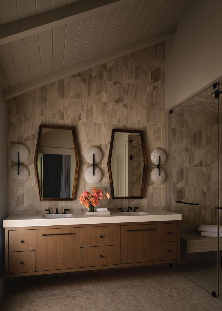 A modern bathroom features a double vanity with wood drawers, two geometric mirrors, and black fixtures. Sconces with globe lights flank each mirror. A vase with pink flowers sits on the white countertop. The walls and floor have beige geometric tiles.