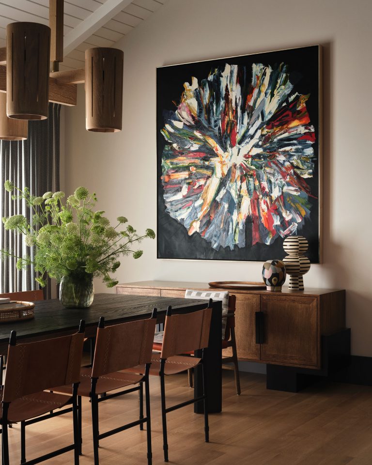 A modern dining room with tan leather chairs, a dark table, and a large abstract painting featuring bold, colorful strokes on a black background. A wooden sideboard holds stacked ceramics and a globe vase; a green leafy plant decorates the table.