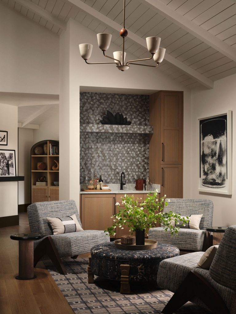 A cozy modern living room with gray armchairs, a round ottoman, and a green plant centerpiece. In the background, a small kitchenette has patterned tile backsplash and wooden cabinets. Artworks and shelves with books decorate the neutral-toned space.
