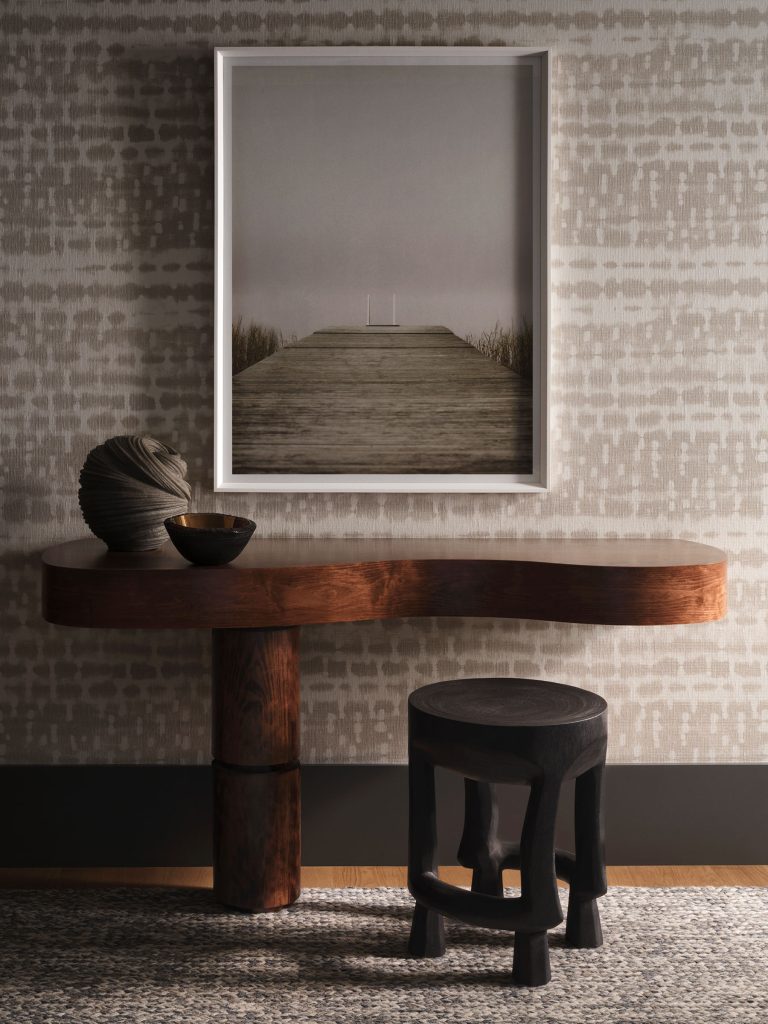 A modern entryway with a curved wooden console table holding a round ceramic sculpture and a small bowl. Above, a framed photo of a pier or boardwalk hangs on a textured gray wall. A black, sculptural stool sits on a woven rug in front of the table.