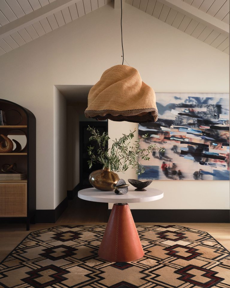 A modern entryway features a round white table with a brown cone base, decorated with a gold vase holding leafy branches and two smaller bowls. Above hangs a woven pendant light. A geometric rug covers the floor, and an abstract painting hangs on the wall behind.