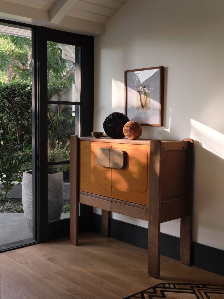 Sunlight streams through glass doors onto a wooden sideboard with tan leather panels. On top are two decorative vases, one black and one brown. A framed photo hangs above. Outside, greenery and a large planter are visible. The floor is pale wood.