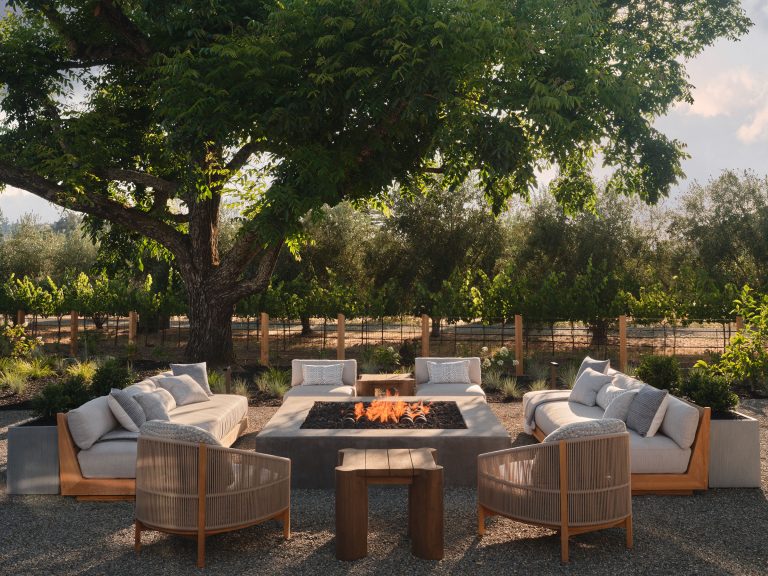 A cozy outdoor seating area with cushioned sofas and chairs surrounds a rectangular fire pit. The space is shaded by a large leafy tree and overlooks a lush garden and wooden fence, with sunlight filtering through green foliage in the background.