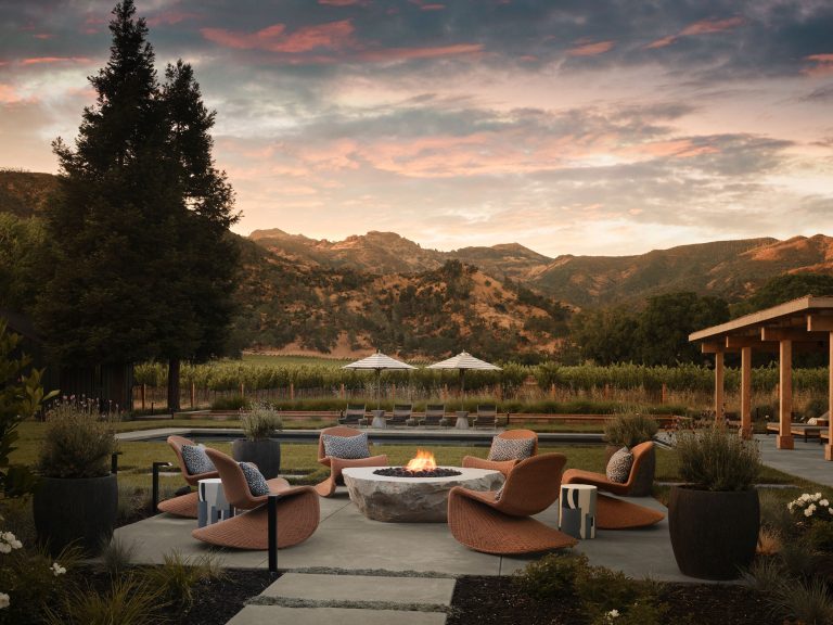 A circular outdoor fire pit with modern chairs sits on a patio surrounded by potted plants, overlooking a vineyard and rolling mountains at sunset. Large trees and a wooden pergola are nearby, with umbrellas and lounge chairs in the background under a colorful sky.