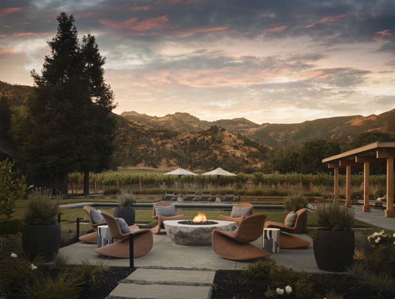 Modern outdoor lounge with curved wicker chairs and cushions arranged around a stone fire pit. Large planters line the patio. Vineyard, umbrellas, and wooden pavilion in background. Mountains and a colorful sunset sky complete the scenic view.