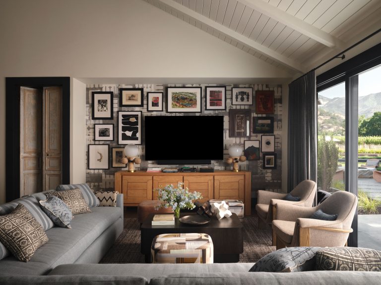 A modern living room with gray couches, patterned pillows, a wooden console, and a flat-screen TV. The wall behind the TV features a grid of framed art. Two beige armchairs and a dark coffee table sit on a textured rug. Large windows reveal scenic outdoor views.