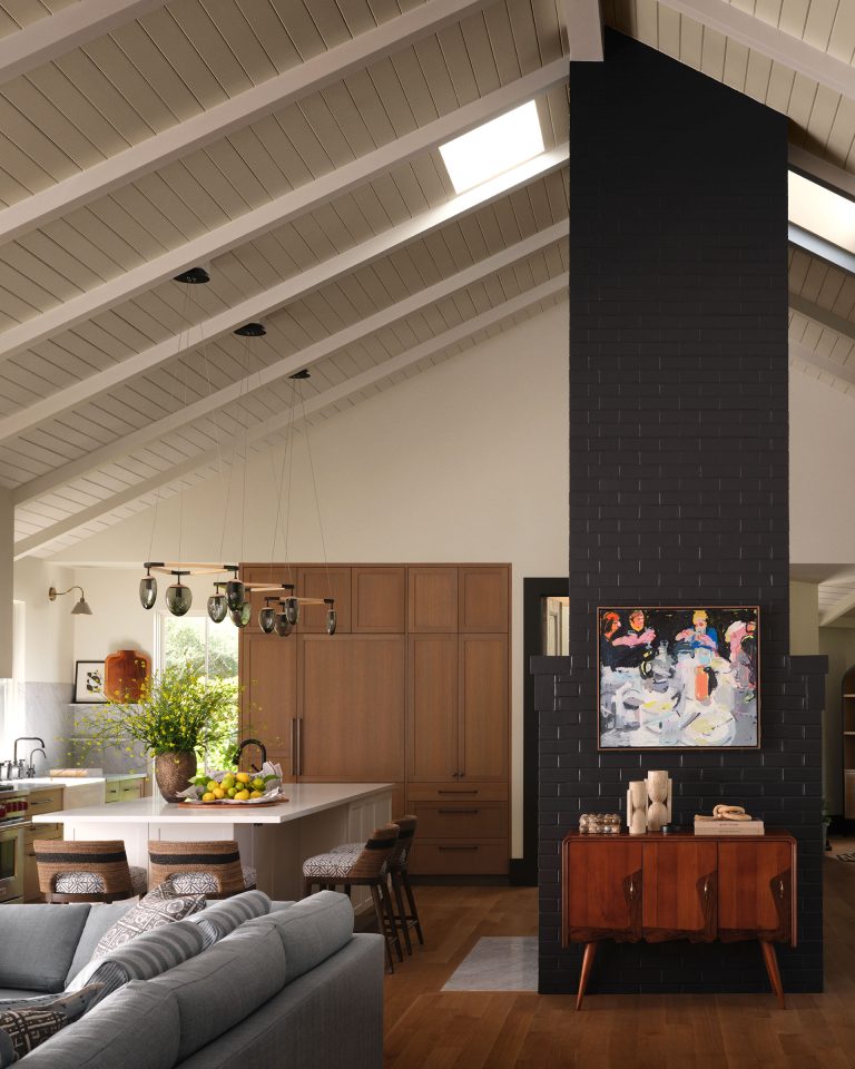 A modern kitchen with vaulted white ceilings and exposed beams features wood cabinets, a central island with stools, hanging pendant lights, and leafy plants. A dark brick chimney with colorful art divides the space; a blue sofa and wooden sideboard sit in the foreground.